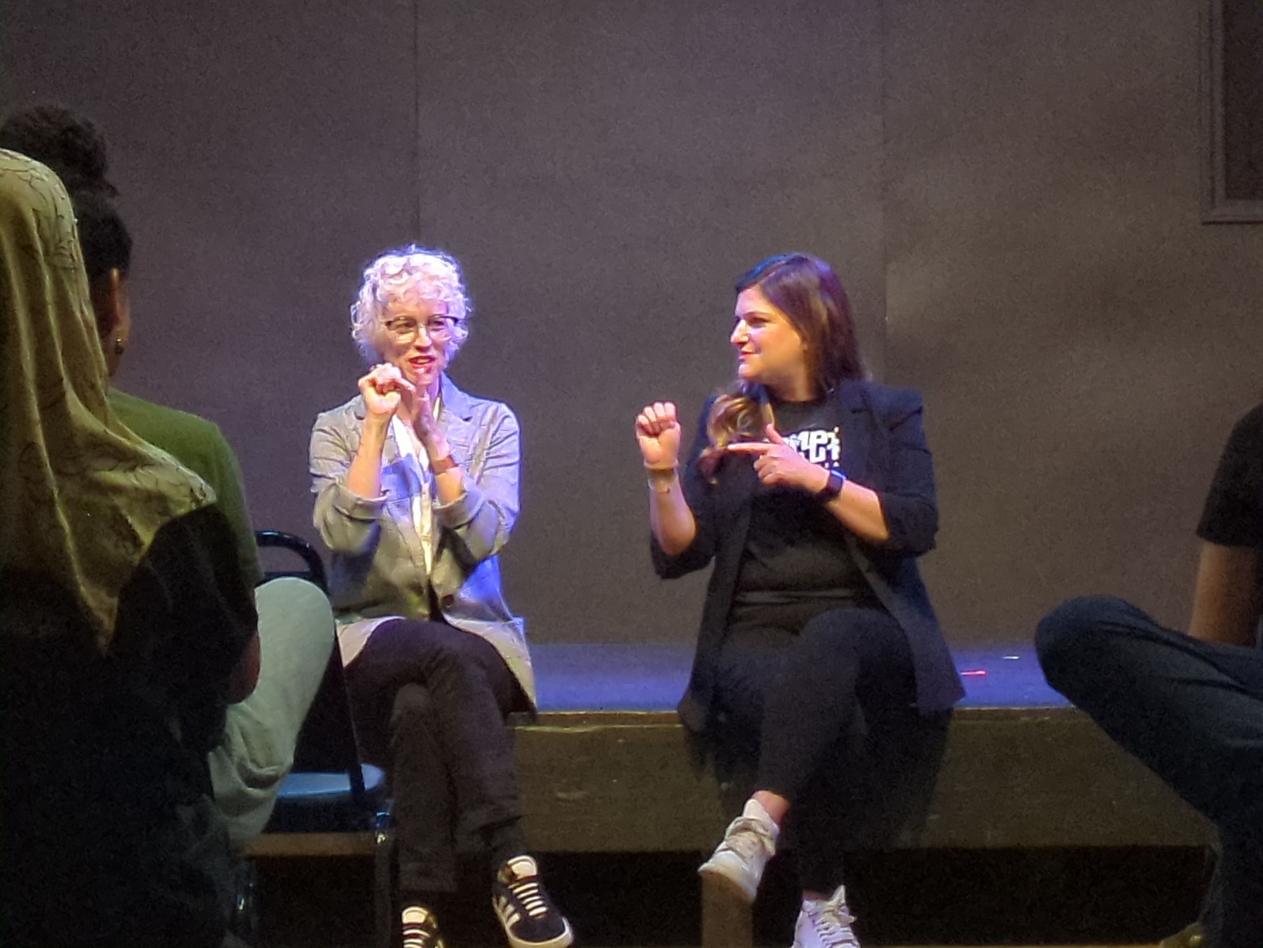 Two women sitting on the edge of a stage gesturing with their hands.