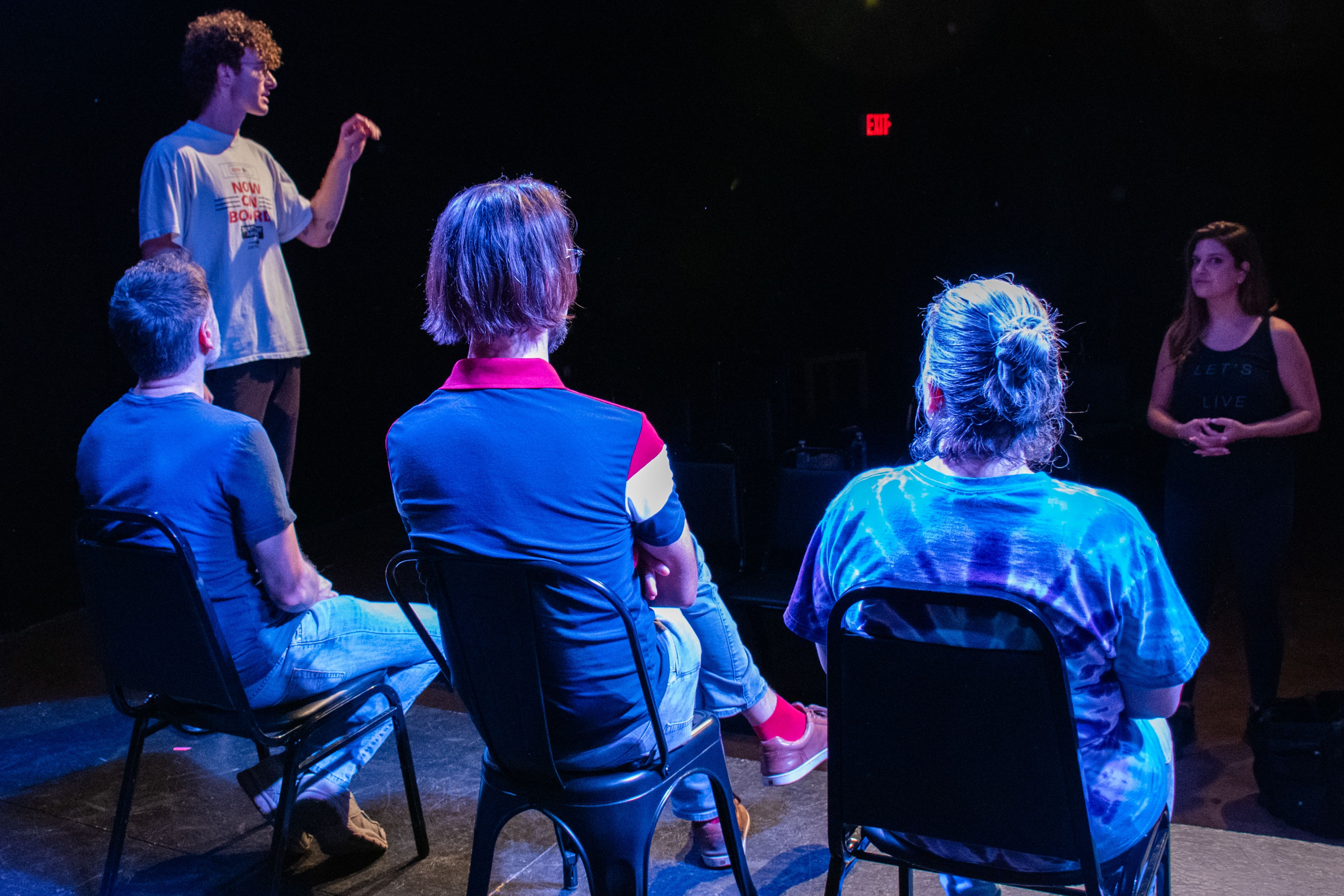 Three people sit on a theater stage with the backs to the camera. Two instructors talk in a darkened theater. 