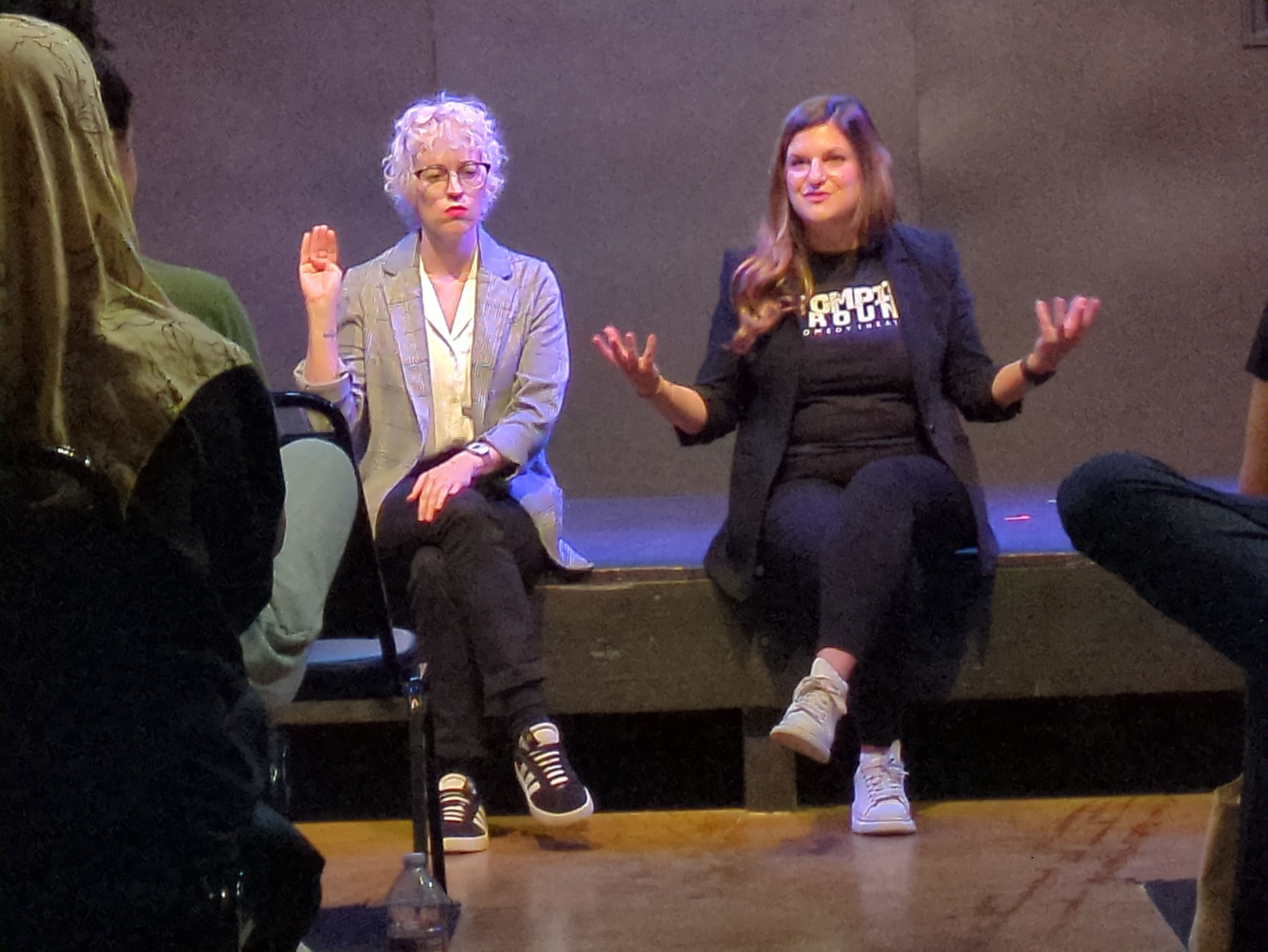 Two women sitting on the edge of a stage gesturing with their hands.