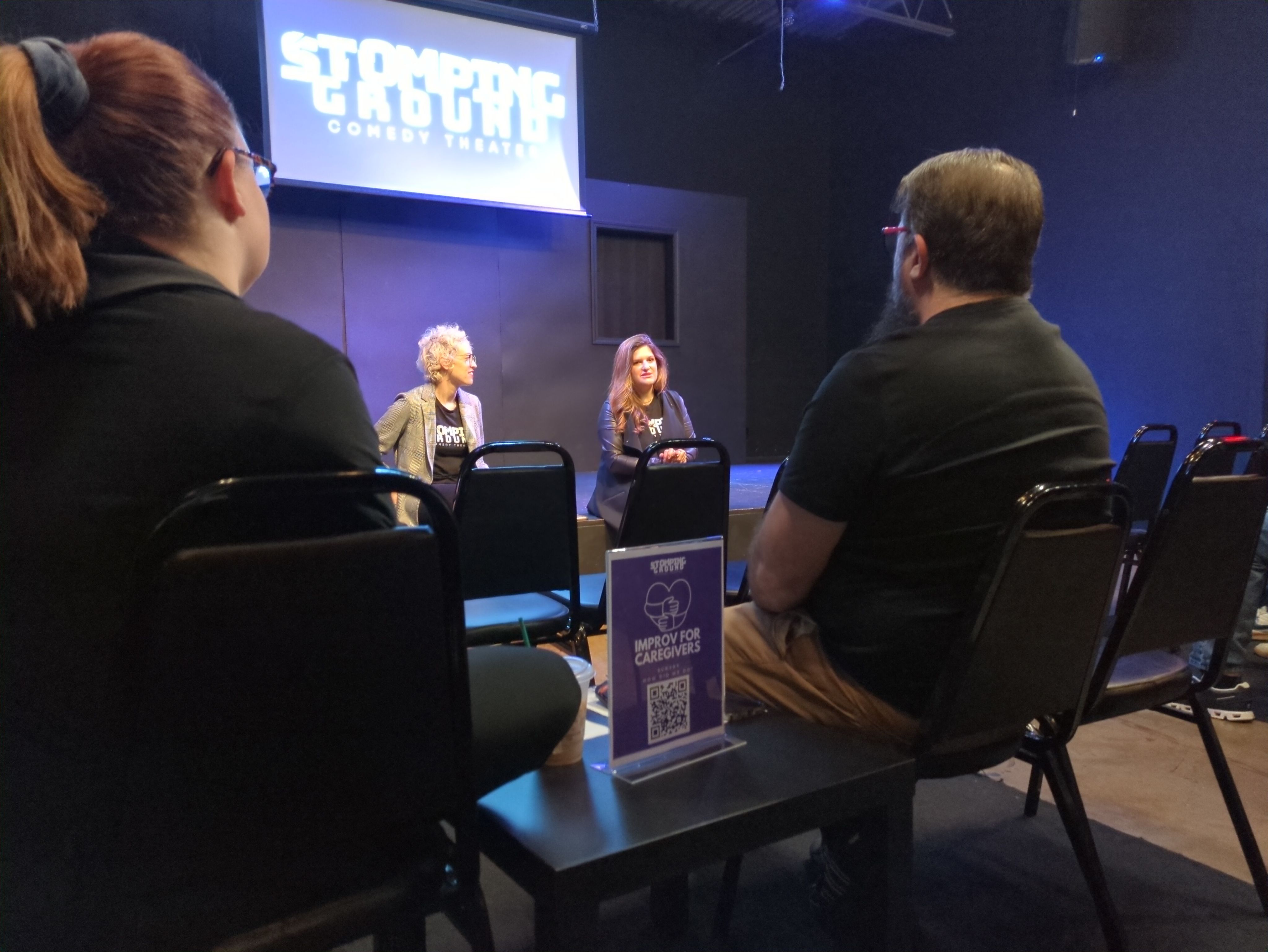 Two women sit on the edge of a theater stage teaching a class.