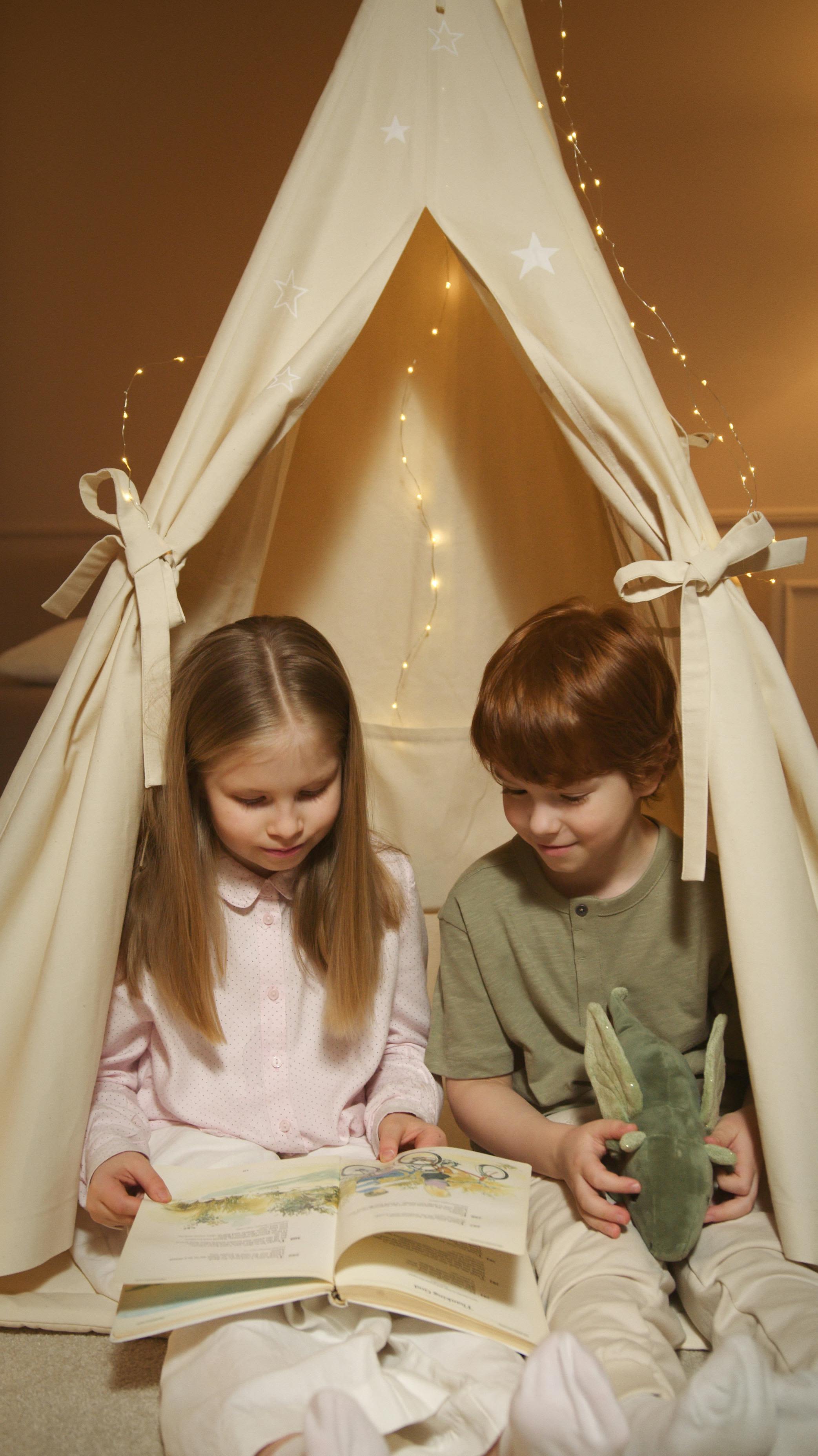 Two kids enjoy story time in a cozy indoor tent with warm lighting.